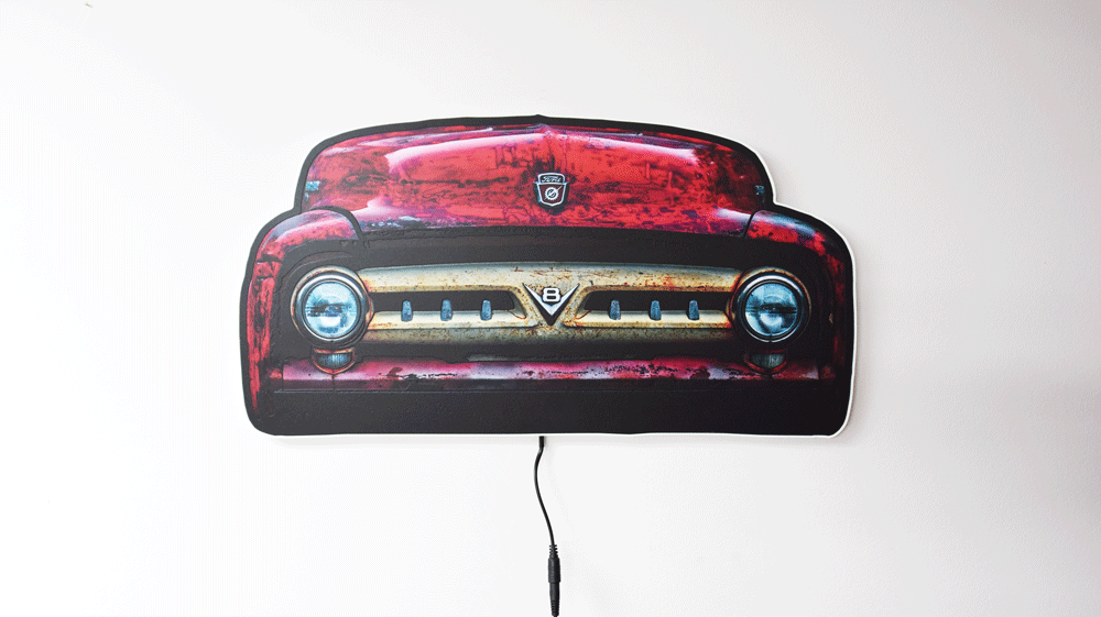 A vintage 1953 F100 grill picture we took and turned it into wall light. The light is off highlighting the distresses red body color and off white center grill with the V8 emblem and Ford hood emblem of the era hanging on a gray wall with a power cord visible below.