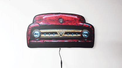 A vintage 1953 F100 grill picture we took and turned it into wall light. The light is off highlighting the distresses red body color and off white center grill with the V8 emblem and Ford hood emblem of the era hanging on a gray wall with a power cord visible below.