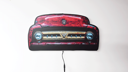 A vintage 1953 F100 grill picture we took and turned it into wall light. The light is off highlighting the distresses red body color and off white center grill with the V8 emblem and Ford hood emblem of the era hanging on a gray wall with a power cord visible below.