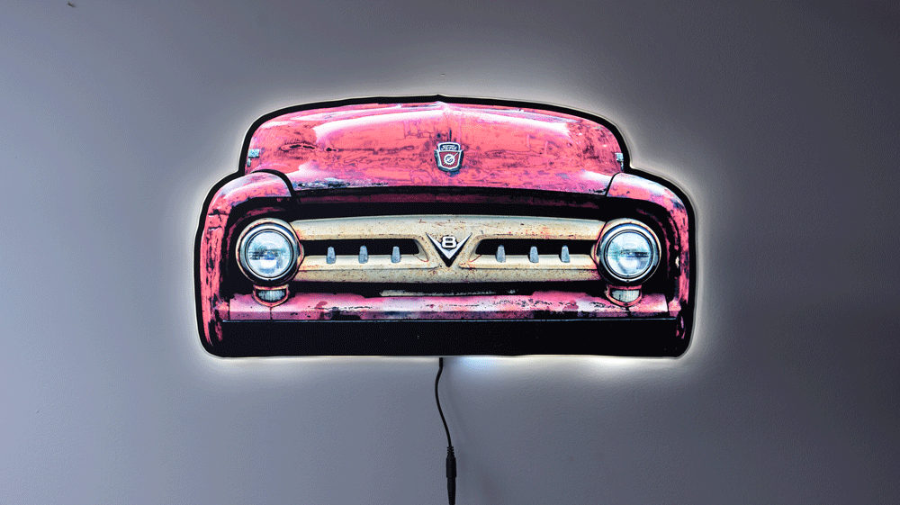 A vintage 1953 F100 grill picture we took and turned it into wall light. The light is on highlighting the distresses red body color and off white center grill with the V8 emblem and ford hood emblem of the era hanging on a gray wall glowing with a power cord visible below.
