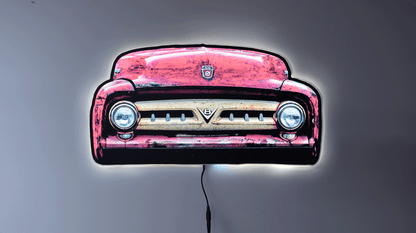 A vintage 1953 F100 grill picture we took and turned it into wall light. The light is on highlighting the distresses red body color and off white center grill with the V8 emblem and ford hood emblem of the era hanging on a gray wall glowing with a power cord visible below.