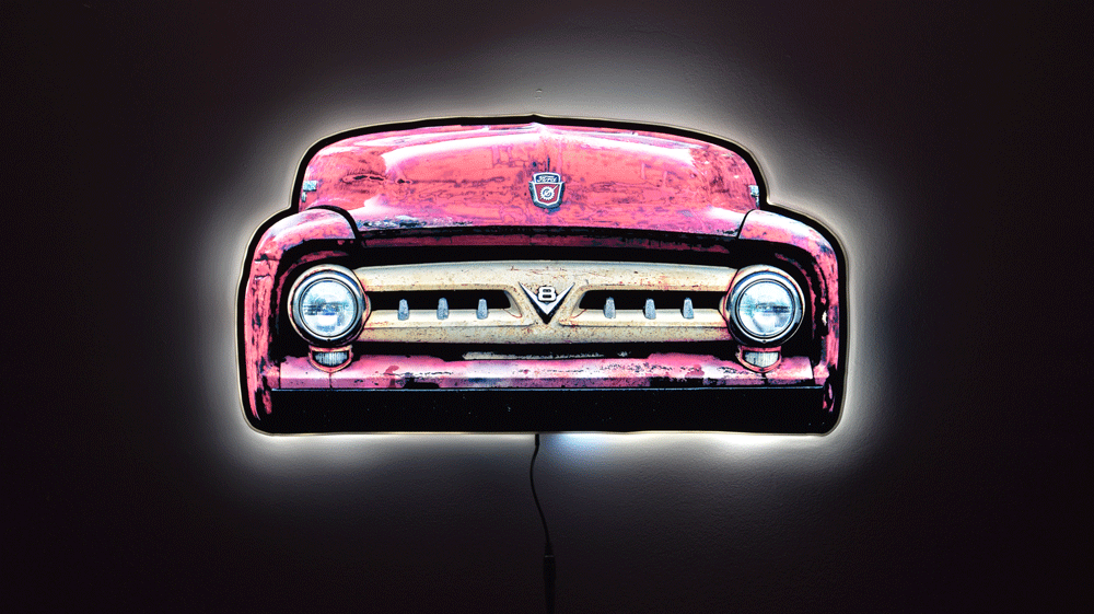 A 1953 F100 front grill wall light, shown against a dark background with a glowing white outline around the illuminated edges.  Front grille with round headlights and a small Ford crest emblem at the top center and a V8 emblem in the center of the classic toothy truck grille with power cord hanging from the bottom.