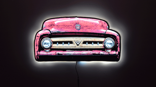 A 1953 F100 front grill wall light, shown against a dark background with a glowing white outline around the illuminated edges.  Front grille with round headlights and a small Ford crest emblem at the top center and a V8 emblem in the center of the classic toothy truck grille with power cord hanging from the bottom.