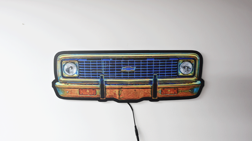 A vintage-1970 K10 front grille wall light with a blue illuminated grille, black bumper outline, and warm orange-brown lighted sections, shown against a plain white wall with a black power cord hanging down.