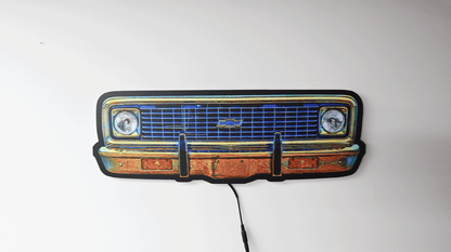 A vintage-1970 K10 front grille wall light with a blue illuminated grille, black bumper outline, and warm orange-brown lighted sections, shown against a plain white wall with a black power cord hanging down.