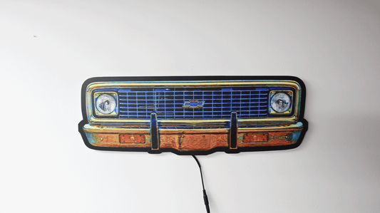 A vintage-1970 K10 front grille wall light with a blue illuminated grille, black bumper outline, and warm orange-brown lighted sections, shown against a plain white wall with a black power cord hanging down.