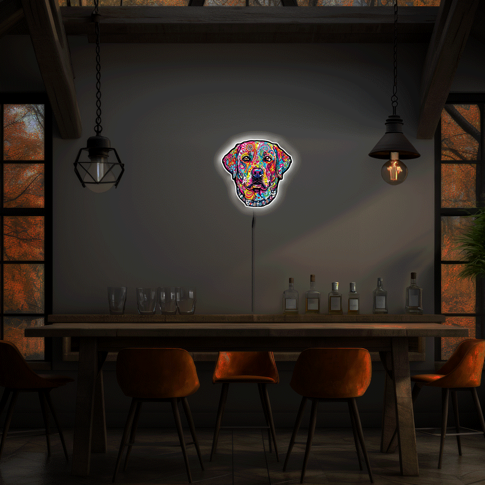 A colorful neon-style wall sign shaped like a English Labrador Retriever head hangs above a wooden table in a dimly lit dining or bar interior with hanging pendant lights, chairs, and bottles.