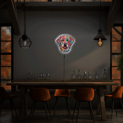 A colorful neon-style wall sign shaped like a English Labrador Retriever head hangs above a wooden table in a dimly lit dining or bar interior with hanging pendant lights, chairs, and bottles.