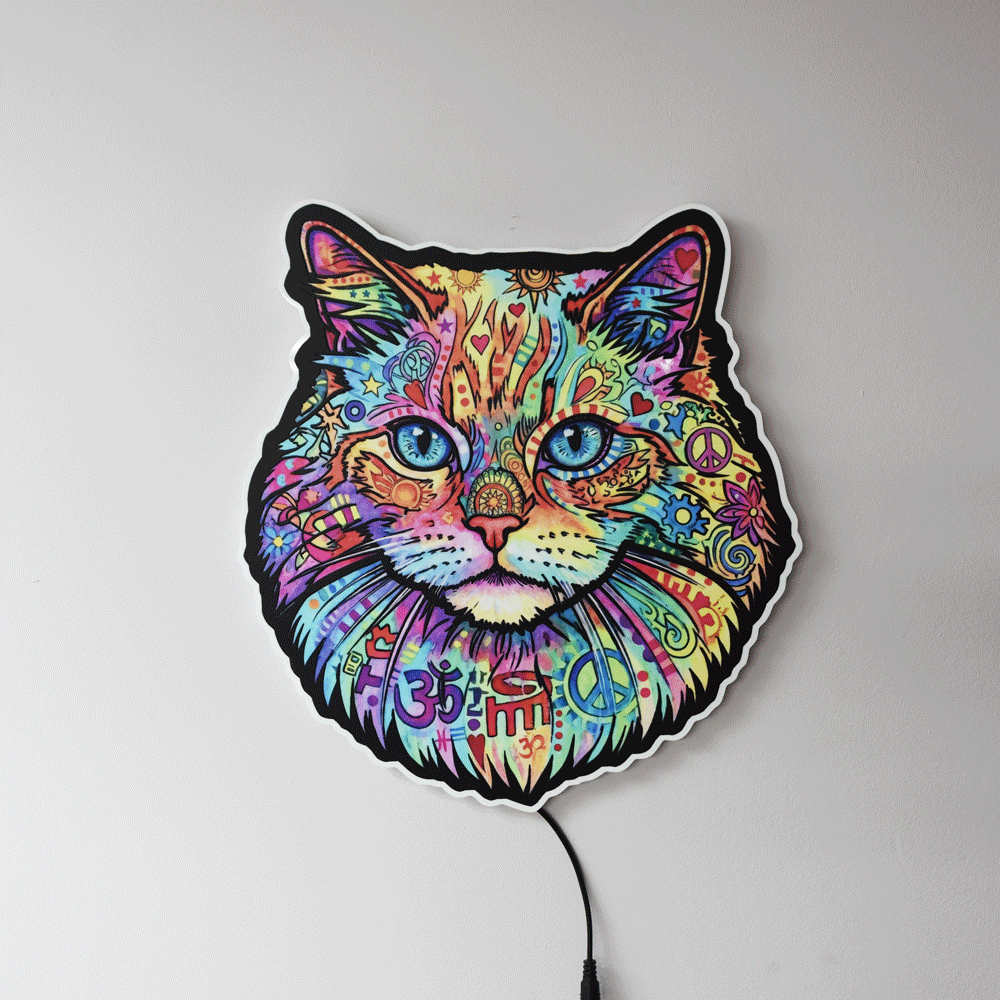 A colorful LED wall light shaped like a cat's face, featuring a vibrant, psychedelic pattern with peace signs, Om symbols, and mandala designs in multiple bright colors. The light is shown mounted on a plain light gray wall with its power cord visible at the bottom.