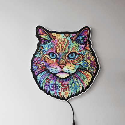 A colorful LED wall light shaped like a cat's face, featuring a vibrant, psychedelic pattern with peace signs, Om symbols, and mandala designs in multiple bright colors. The light is shown mounted on a plain light gray wall with its power cord visible at the bottom.