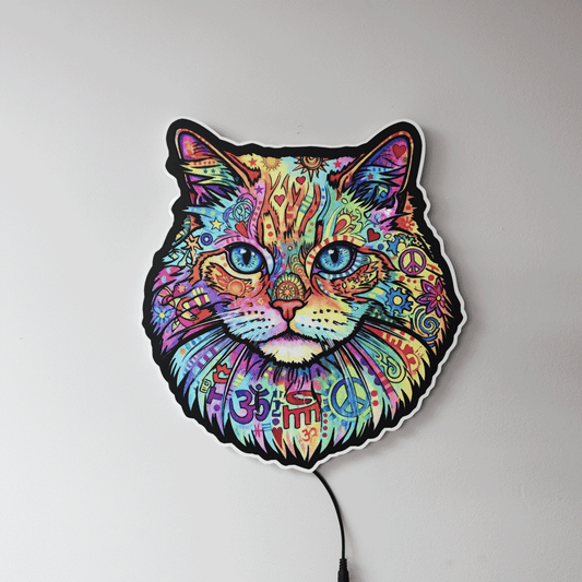 A colorful LED wall light shaped like a cat's face, featuring a vibrant, psychedelic pattern with peace signs, Om symbols, and mandala designs in multiple bright colors. The light is shown mounted on a plain light gray wall with its power cord visible at the bottom.
