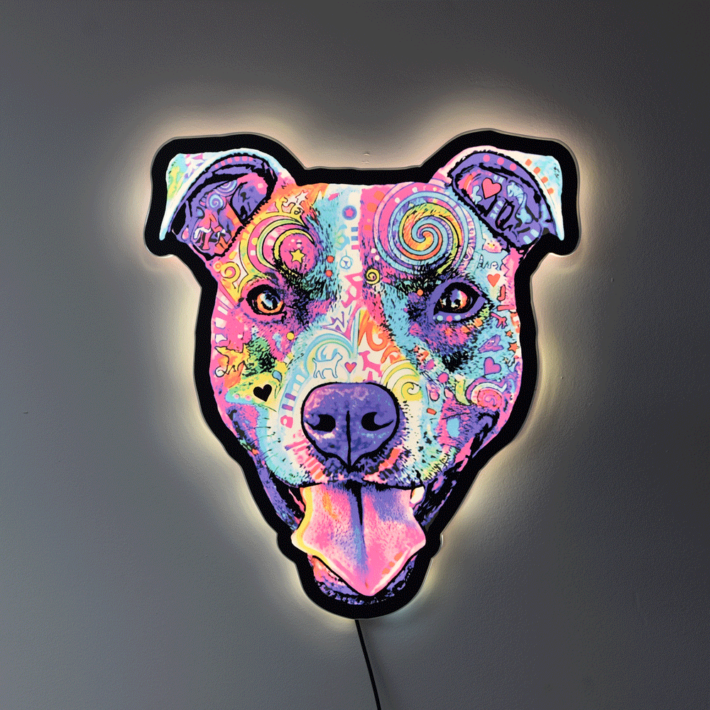 A decorative light shaped like a Colorful Smiling Pit Bull Face with tongue hanging out, featuring a vibrant psychedelic pattern in pink, purple, blue, yellow, and green, with a glowing warm white light around the edges, mounted on a dark gray wall.