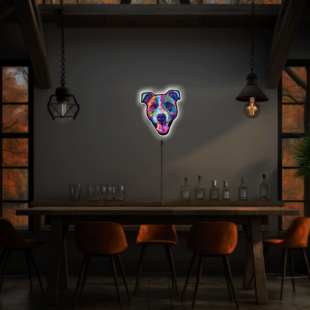 A neon-style wall sign shaped Colorful Smiling Pit Bull Face with tongue hanging out glows above a wooden dining table in a dimly lit modern interior with hanging pendant lights and orange chairs.