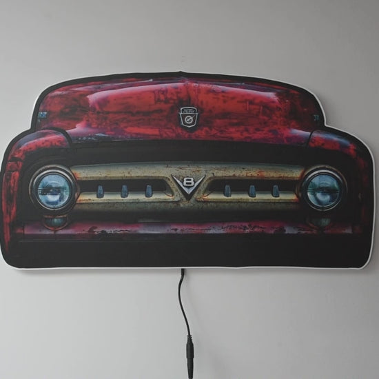 A vintage 1953 F100 grill picture we took and turned it into wall light. The light is off and then on highlighting the distresses red body color and off white center grill with the V8 emblem and ford hood emblem of the era hanging on a gray wall glowing when on with a power cord visible below.