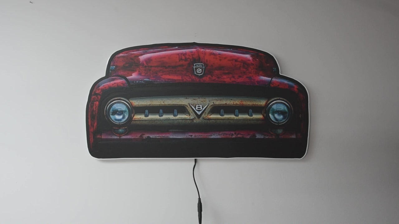 A vintage 1953 F100 grill picture we took and turned it into wall light. The light is off and then on highlighting the distresses red body color and off white center grill with the V8 emblem and ford hood emblem of the era hanging on a gray wall glowing when on with a power cord visible below.