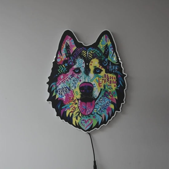 A colorful LED wall light shaped like a wolf or husky head, glowing against a dark gray wall turned on and off in a lighted and dark rooms. The design features a vibrant, multicolored graffiti-style pattern with pink, blue, yellow, and green details, and a black outline.