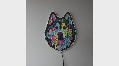 A colorful LED wall light shaped like a wolf or husky head, glowing against a dark gray wall turned on and off in a lighted and dark rooms. The design features a vibrant, multicolored graffiti-style pattern with pink, blue, yellow, and green details, and a black outline.